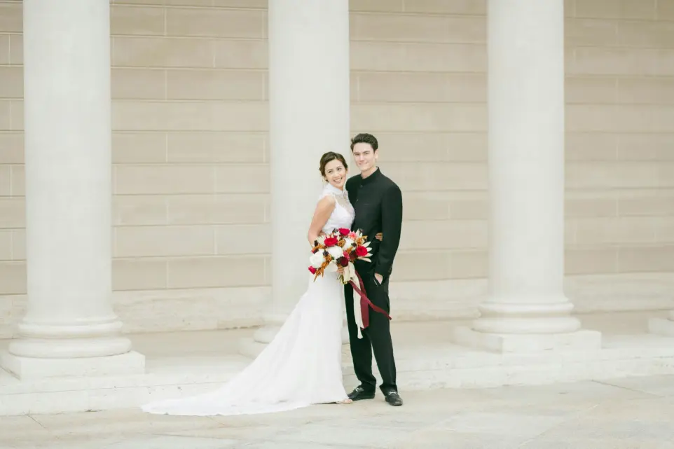 The bride wears a flowing white gown with a lace train, and the groom is in a classic black tuxedo. Both are smiling, radiating happiness and love on their special day.