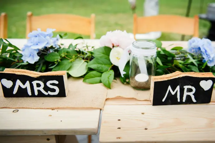Two Hearts, One Soul A beautifully arranged wedding venue featuring wooden stools neatly lined up, each with a guest’s name elegantly written on a tag or calligraphy card. The seating area is decorated with soft florals and ambient lighting, creating a personalized and welcoming atmosphere.